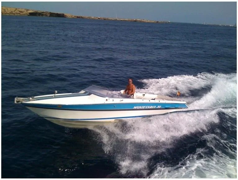 A man is riding a white and blue speedboat on open water, creating a wake behind it during the daytime.