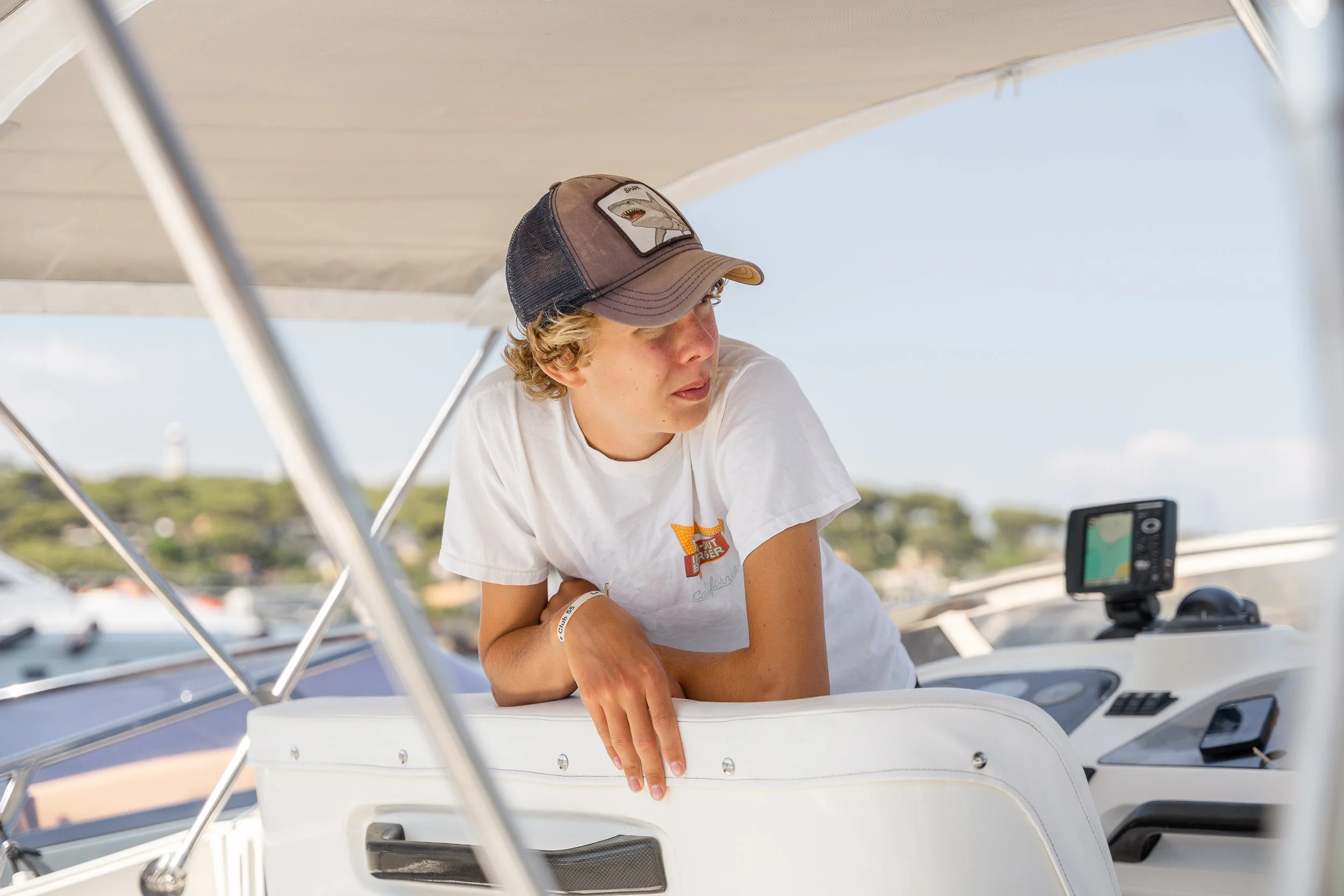 A young person with curly hair, wearing a baseball cap and a white t-shirt, leaning on the helm of a boat, looking off to the side, with navigational equipment and a view of water and land in the background.