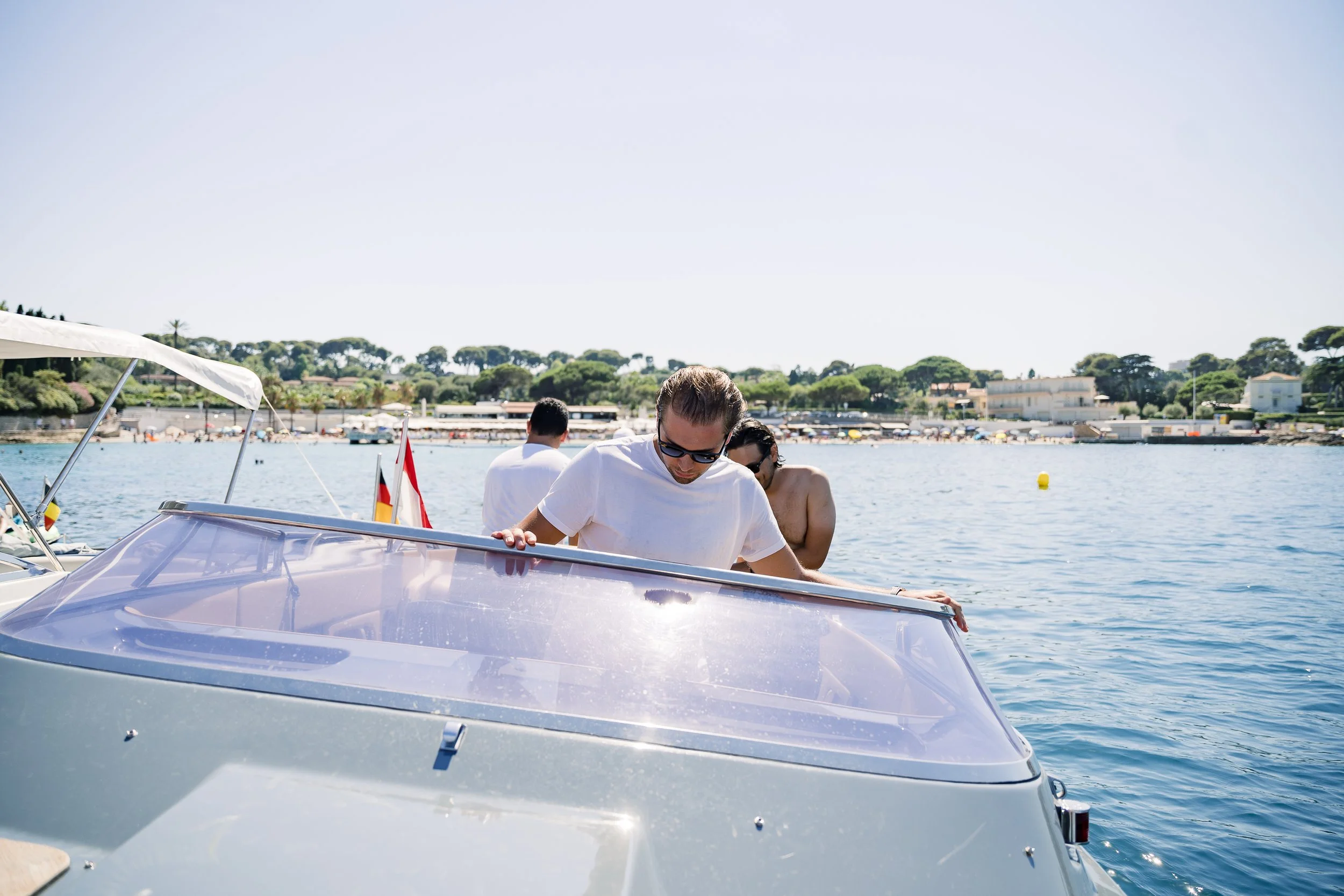 People on a boat by the water, with a man in a white shirt and sunglasses leaning over the boat's windshield, and others in swimwear nearby, a shoreline with trees and buildings in the background.