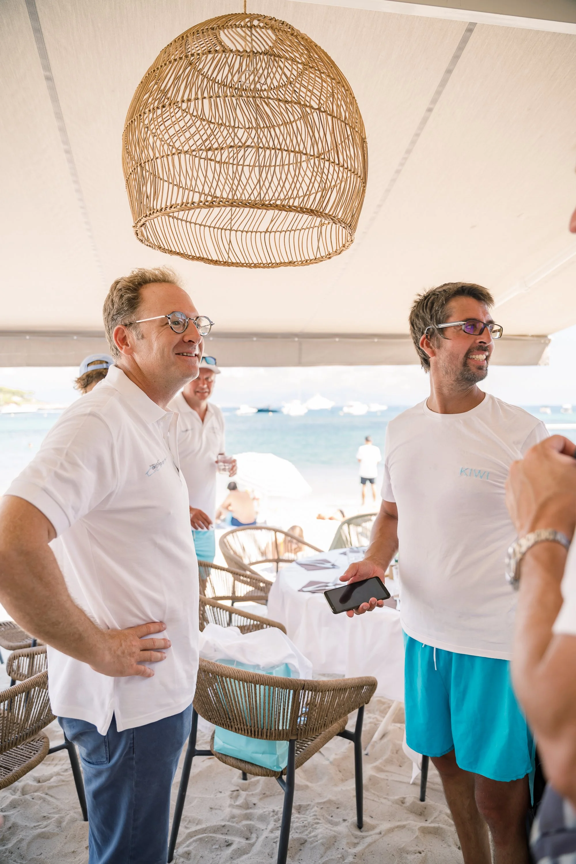 Three men in casual shirts and shorts are standing and talking on a sandy beach patio with tables and chairs, overlooking the ocean with boats in the background.