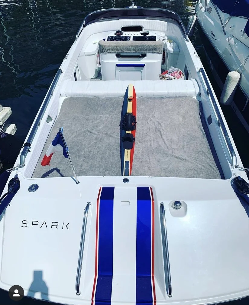 A white motorboat titled 'SPARK' docked at a marina with a folded sail and a small French flag, with water and adjacent boats visible around it.