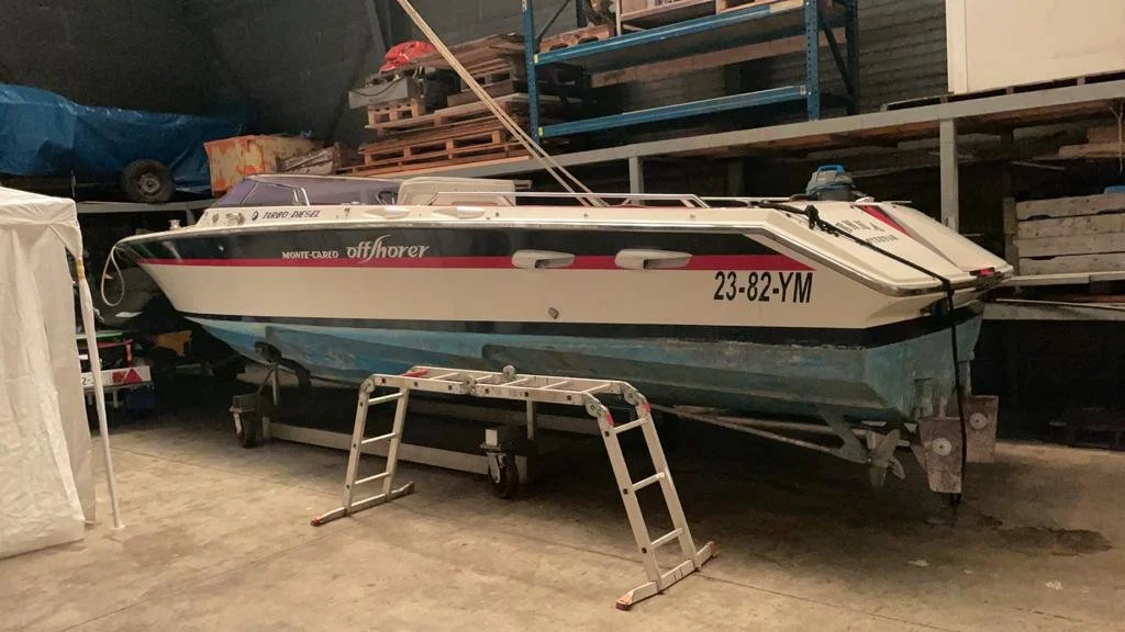 A speedboat with registration 23-82-YM stored indoors on a trailer, with a ladder in front, in a garage or warehouse with shelving and other items.