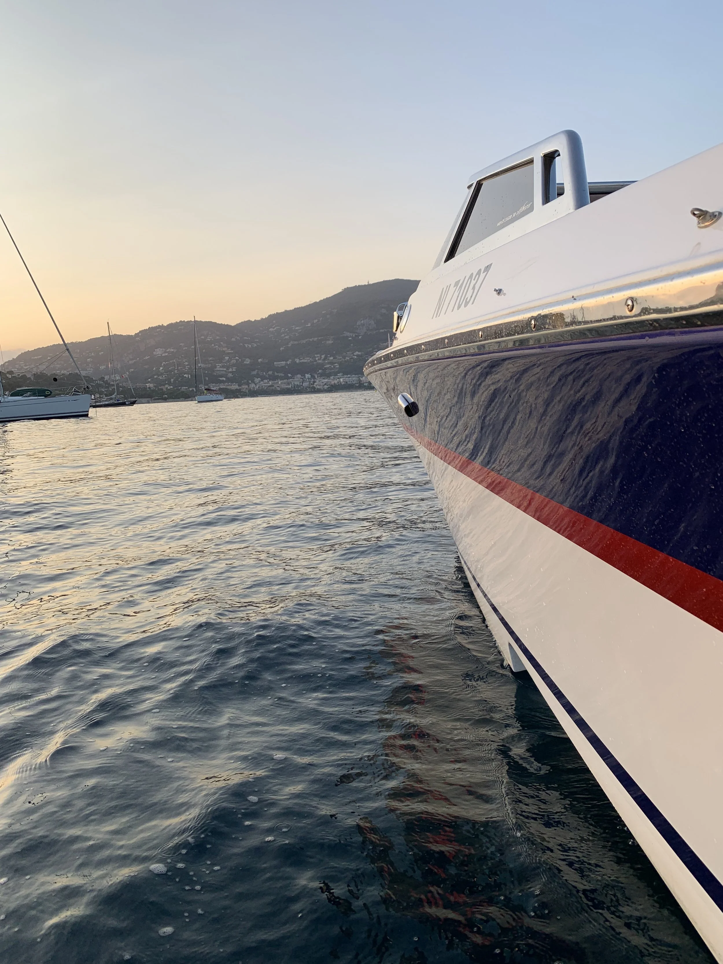 Close-up of a white and blue boat on calm water during sunset with other boats and hills in the background.