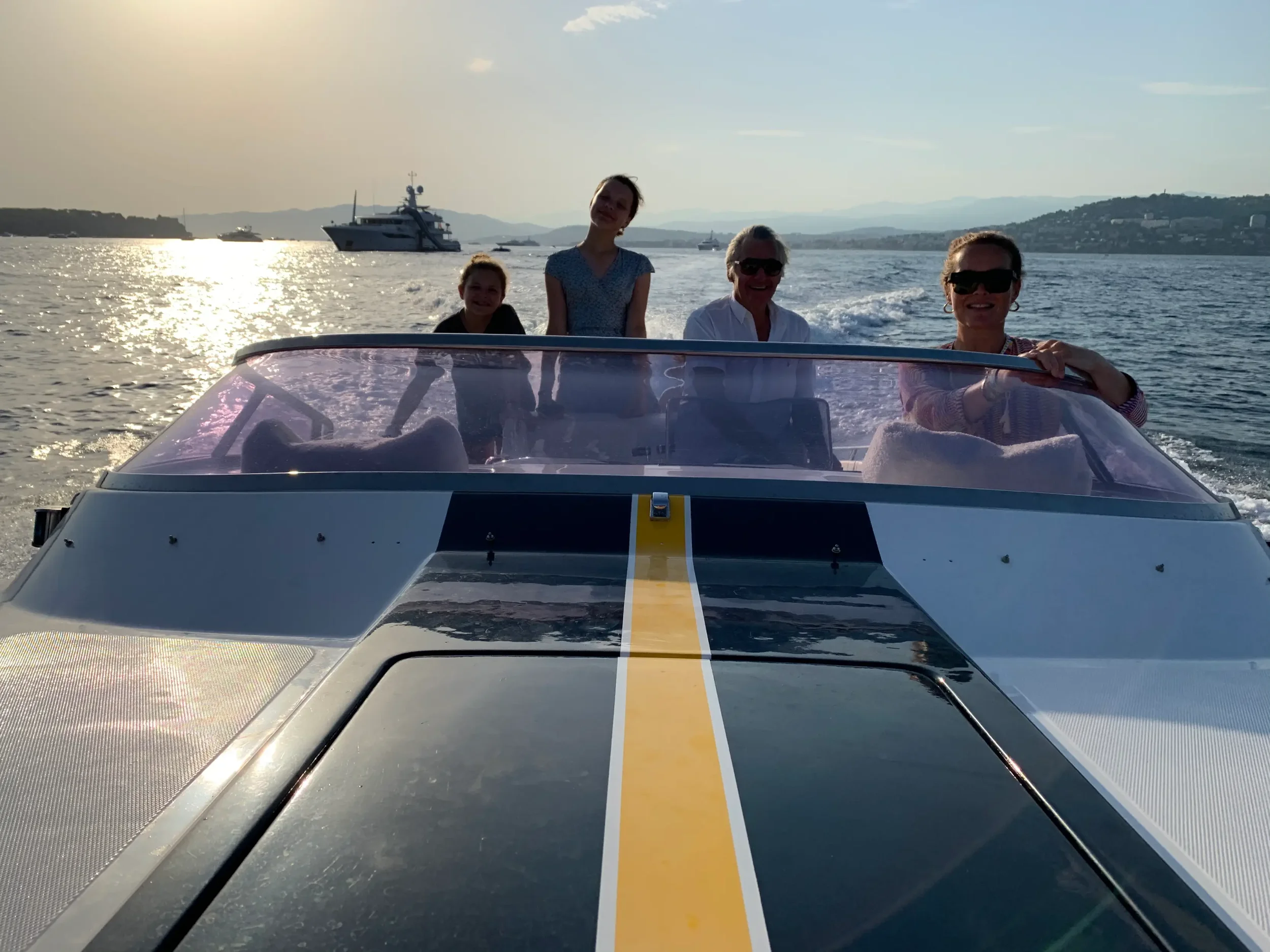 Four people, two children and two adults, are on a boat during sunset, with a waterway and other boats in the background.