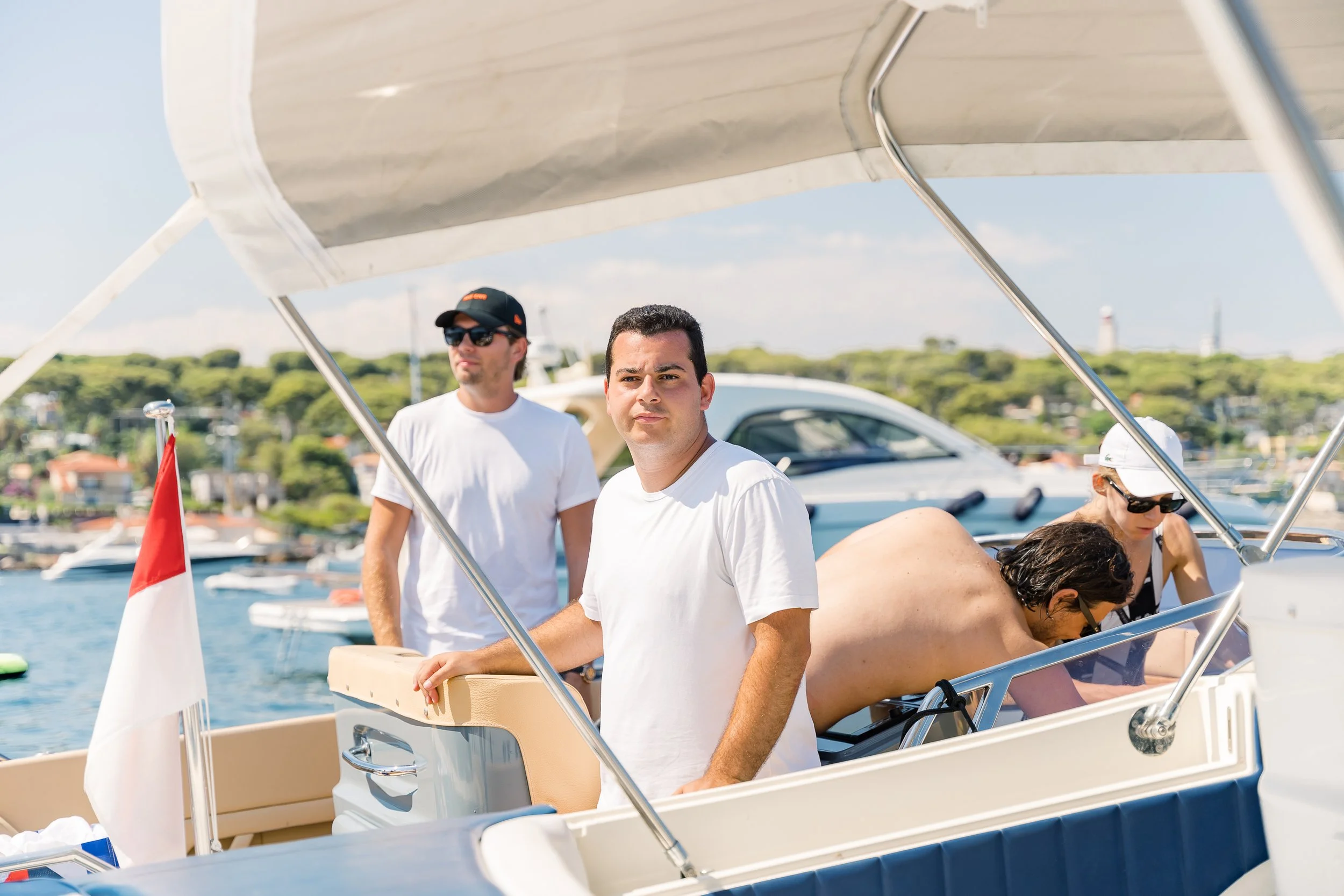 Four people on a boat, three standing and one sitting, with a marina and hills in the background.
