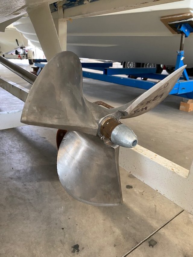 Boat propeller with three blades mounted on its shaft, situated under a boat in a garage or workshop.