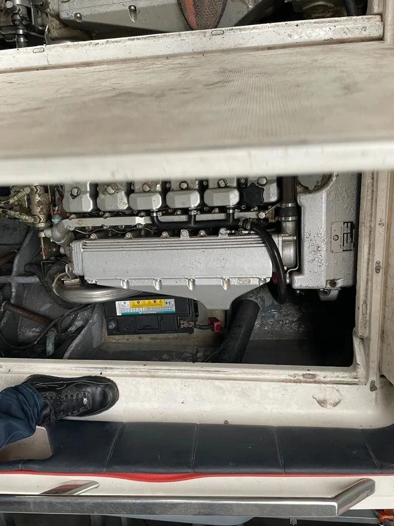 View of a machine engine with visible components including valves, wires, and a battery, seen from above with part of a person's shoe visible at the bottom left.