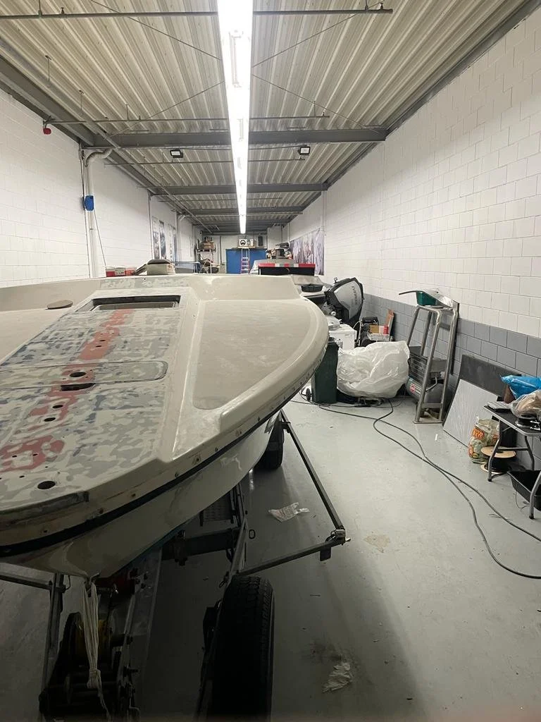 Indoor boat storage with a boat on a trailer, surrounded by tools and equipment, in a garage-like space with white brick walls and a metal ceiling.