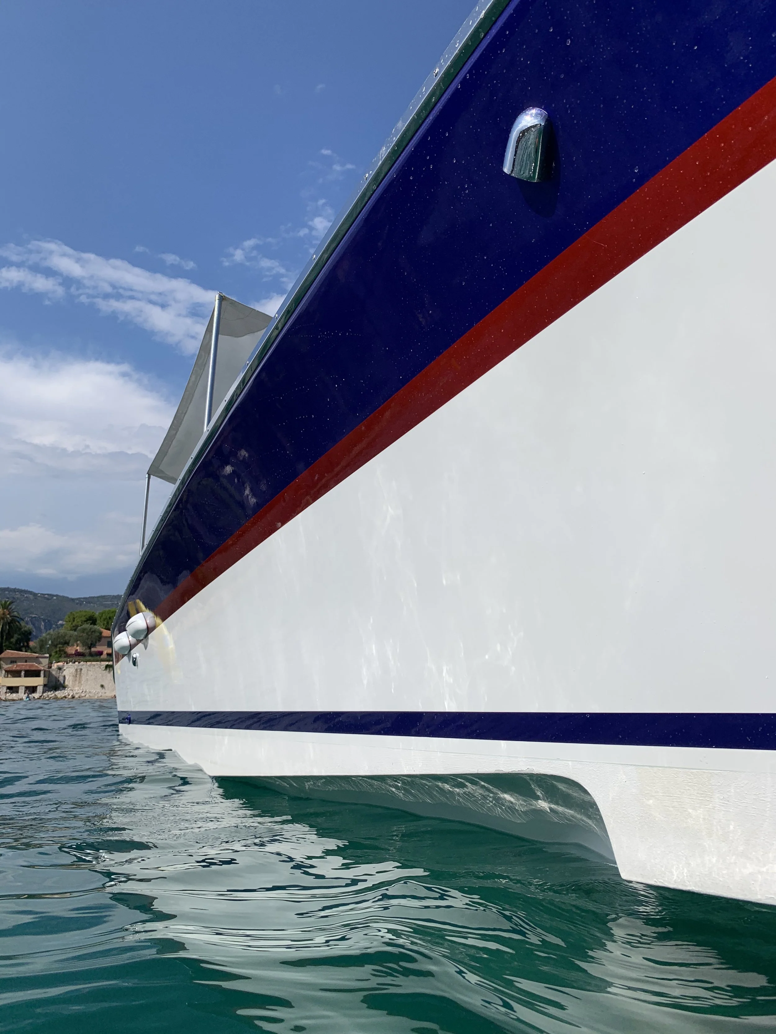 Close-up of a boat's bow with red, white, and blue paint, floating on water near a shoreline with houses and trees under a partly cloudy sky.