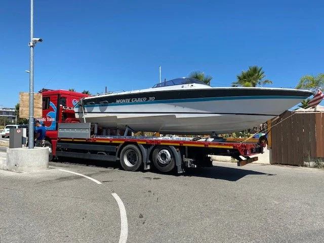 A boat on a flatbed truck in a parking lot with palm trees and a clear blue sky in the background.