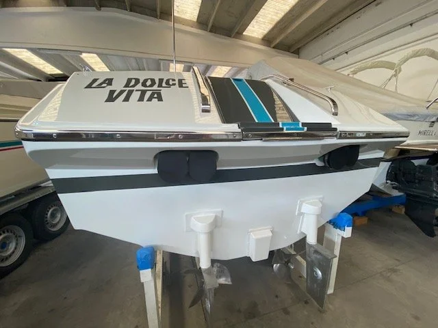 A white speedboat with blue and black stripes and the words 'La Dolce Vita' on the bow, stored indoors on a trailer.