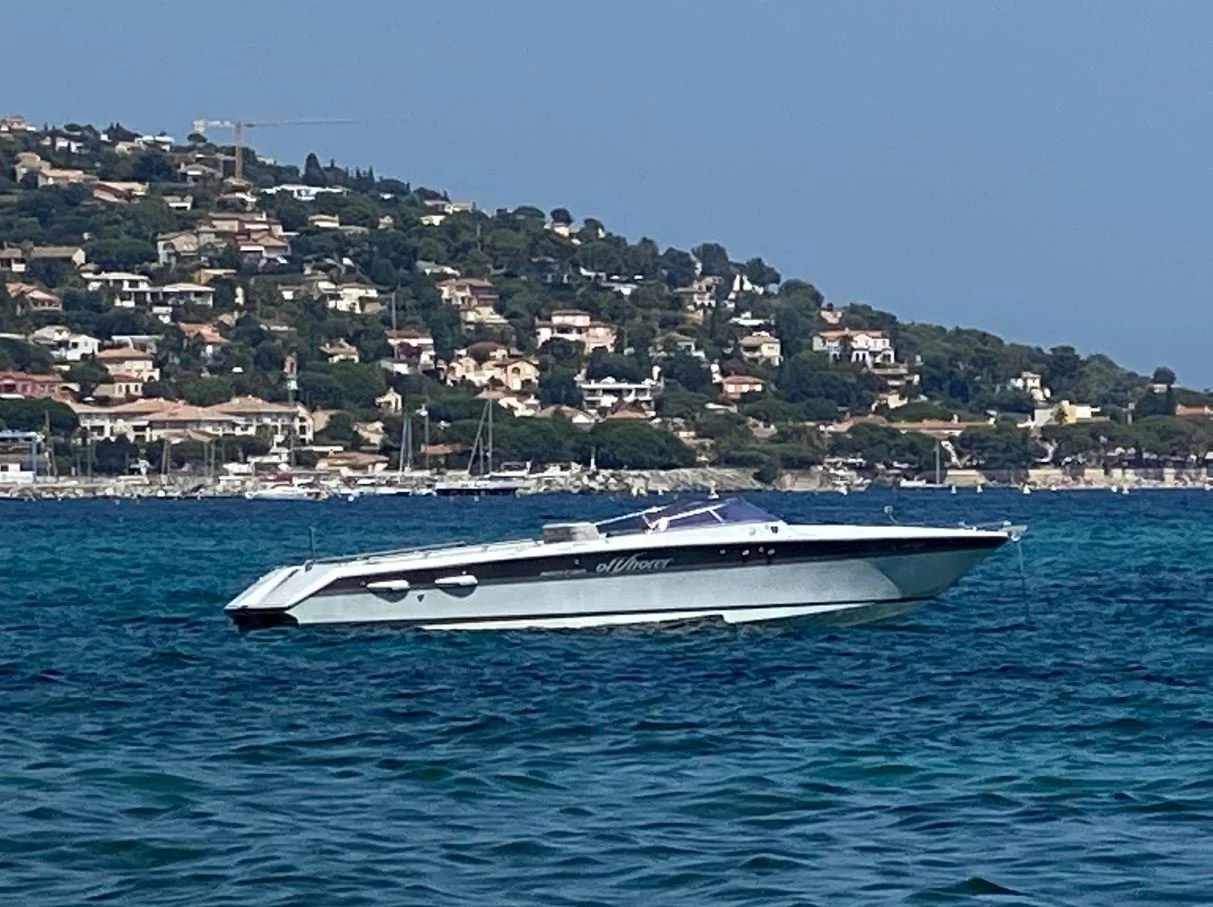 Luxury motor yacht floating on blue water with a hillside residential area and marina in the background under a clear sky.
