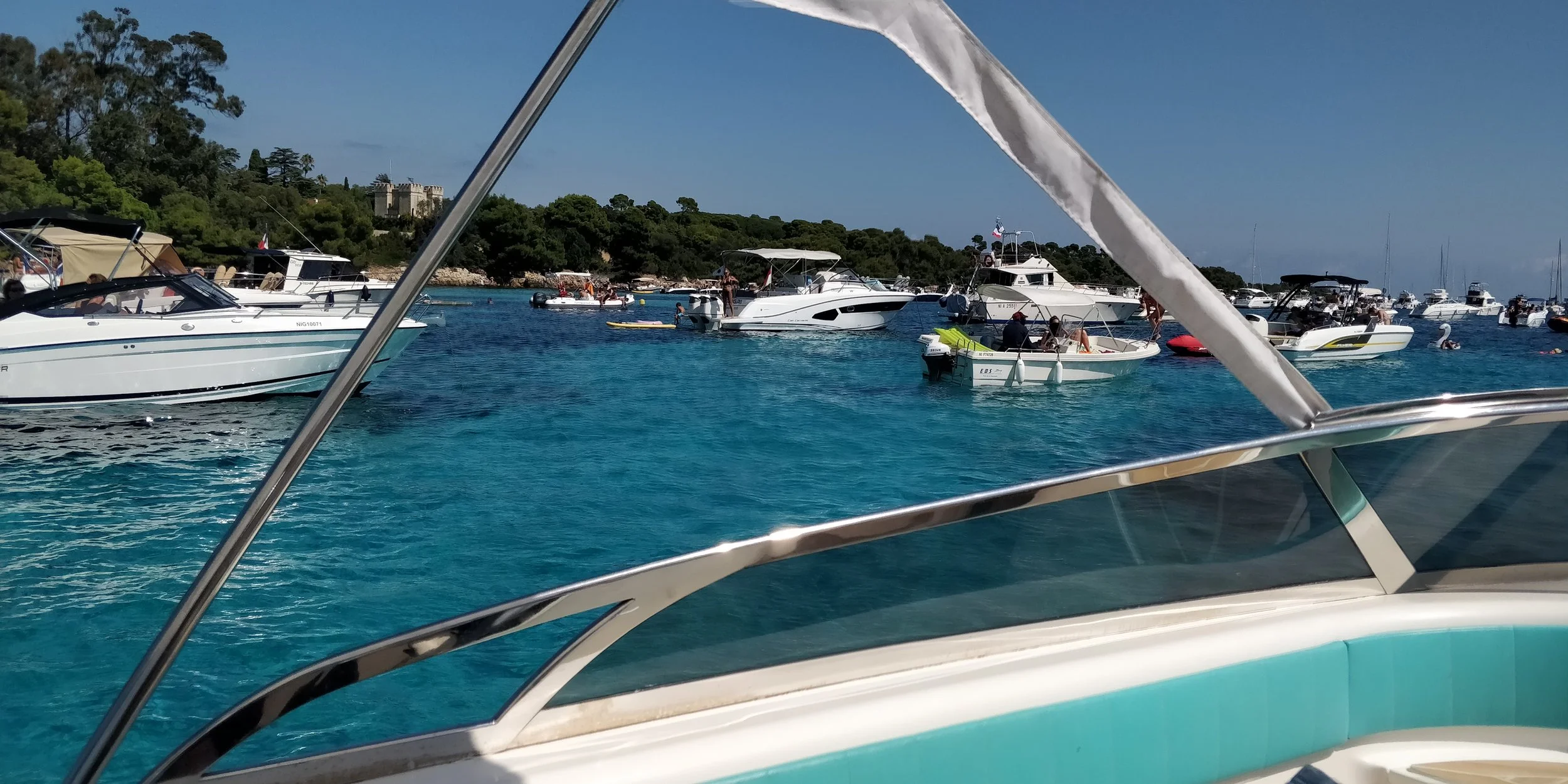 Multiple boats anchored on clear blue water near a tree-lined shoreline under a clear sky.