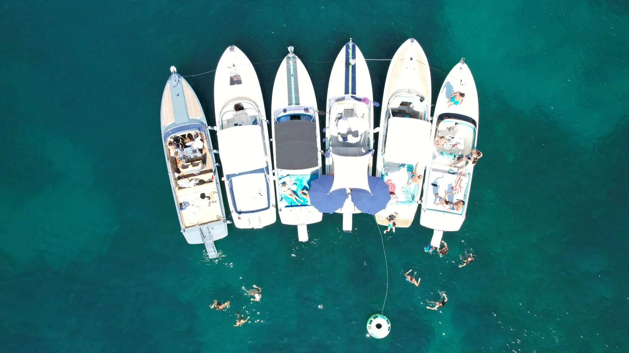 Top-down aerial view of seven boats anchored in the water with people swimming and relaxing on the boats and in the water.