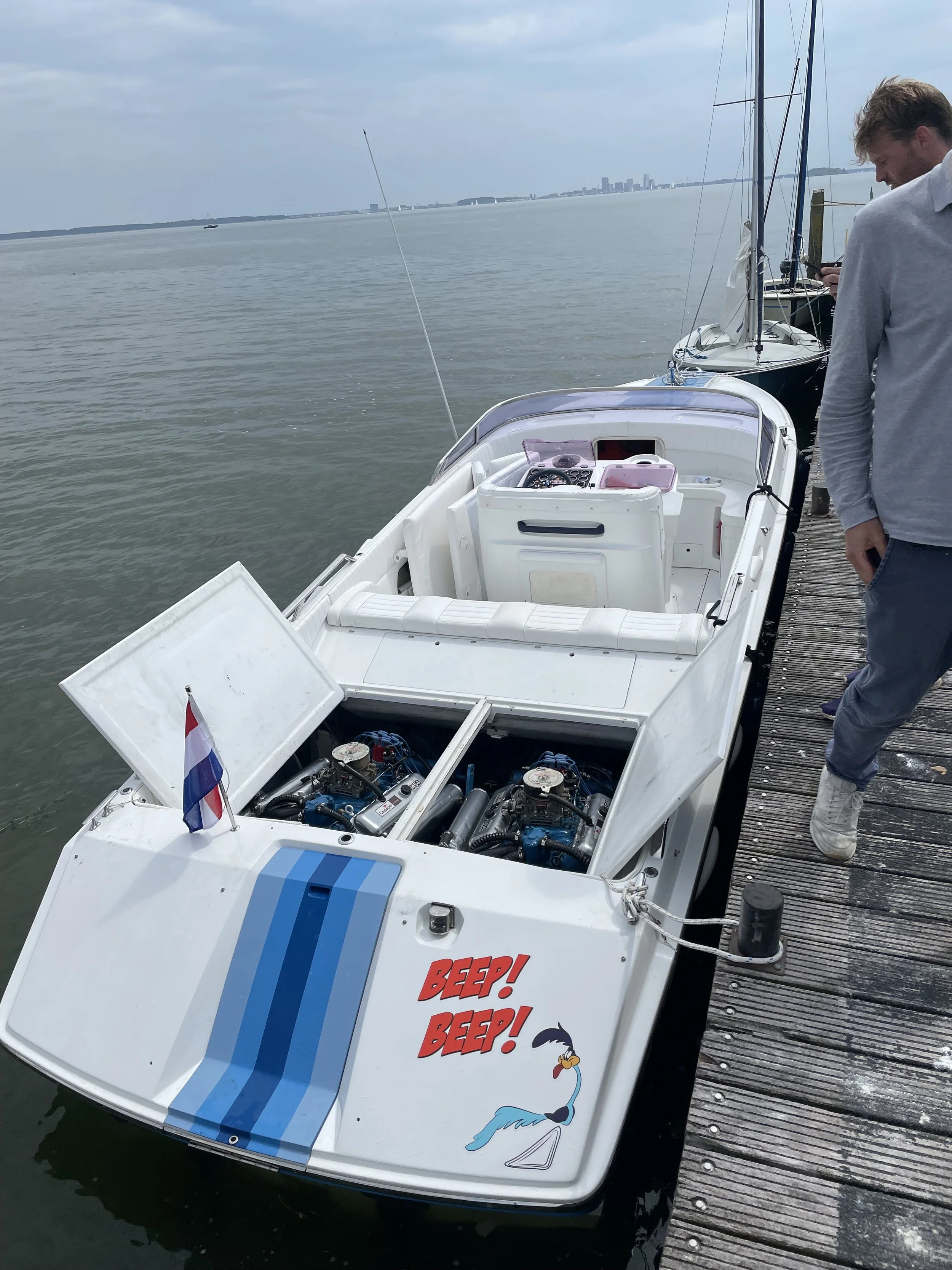 White speedboat docked at a marina with two blue racing stripes, a small Dutch flag, a cartoon cartoon of a stork with the words 'BEEP! BEEP!', and two outboard engines. A person is standing on the dock nearby.