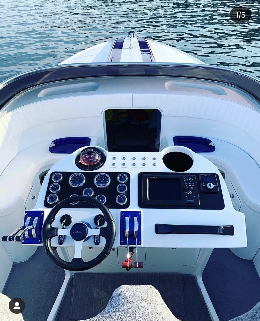 View from the helm of a yacht or boat showing a ship's steering wheel, gauge cluster, navigation system, and seating area, with water in the background.