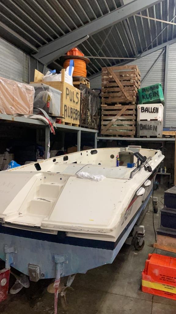 A small white boat on a trailer in a storage warehouse, surrounded by boxes, pallets, and shelves with various items.