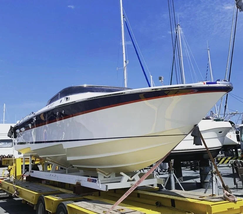 A large white and black motor yacht on a yellow flatbed trailer at a marina, with sailboats and blue sky in the background.
