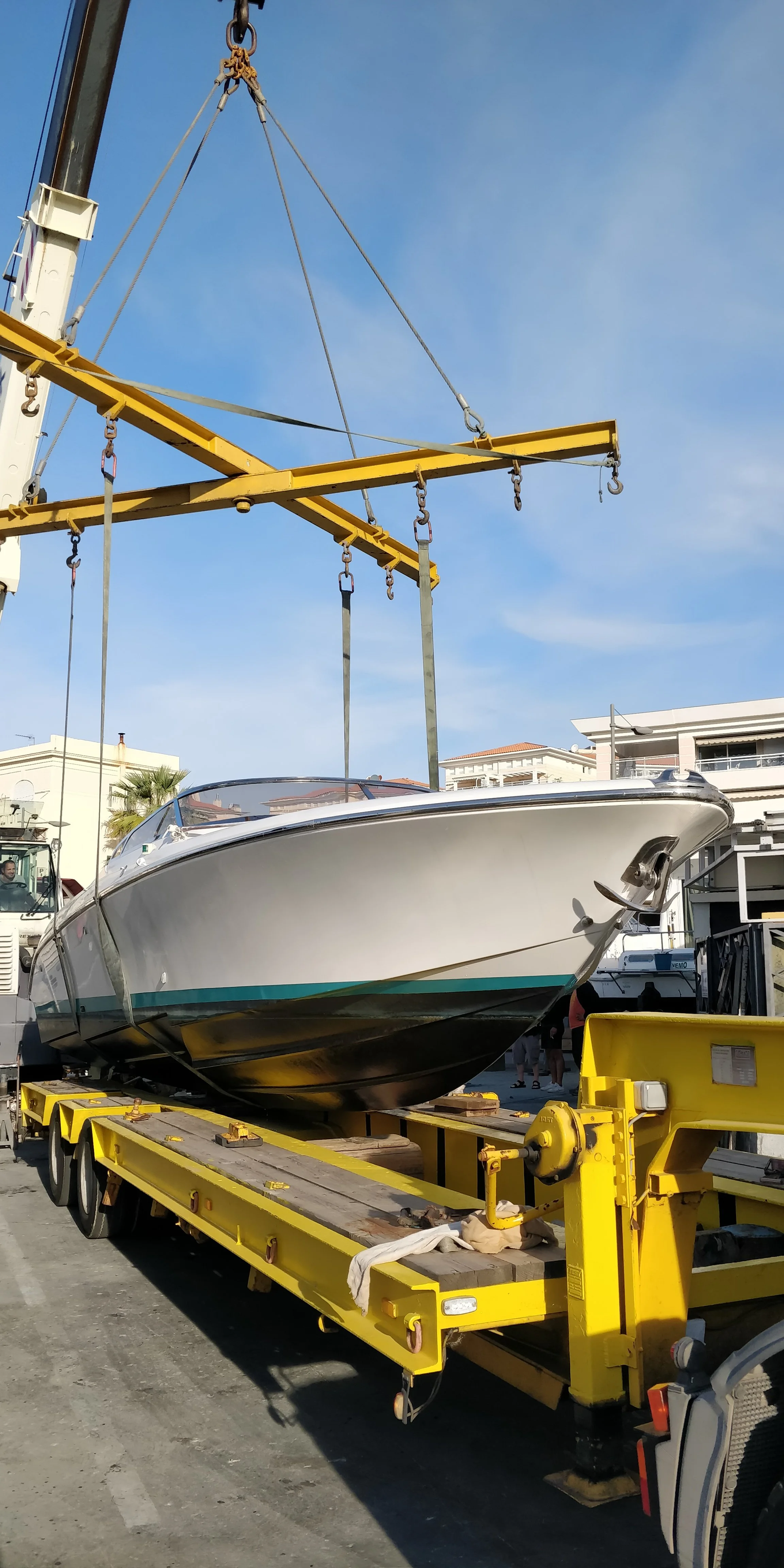 A white boat is being lifted by a yellow crane on a truck bed at a marina or dock area.