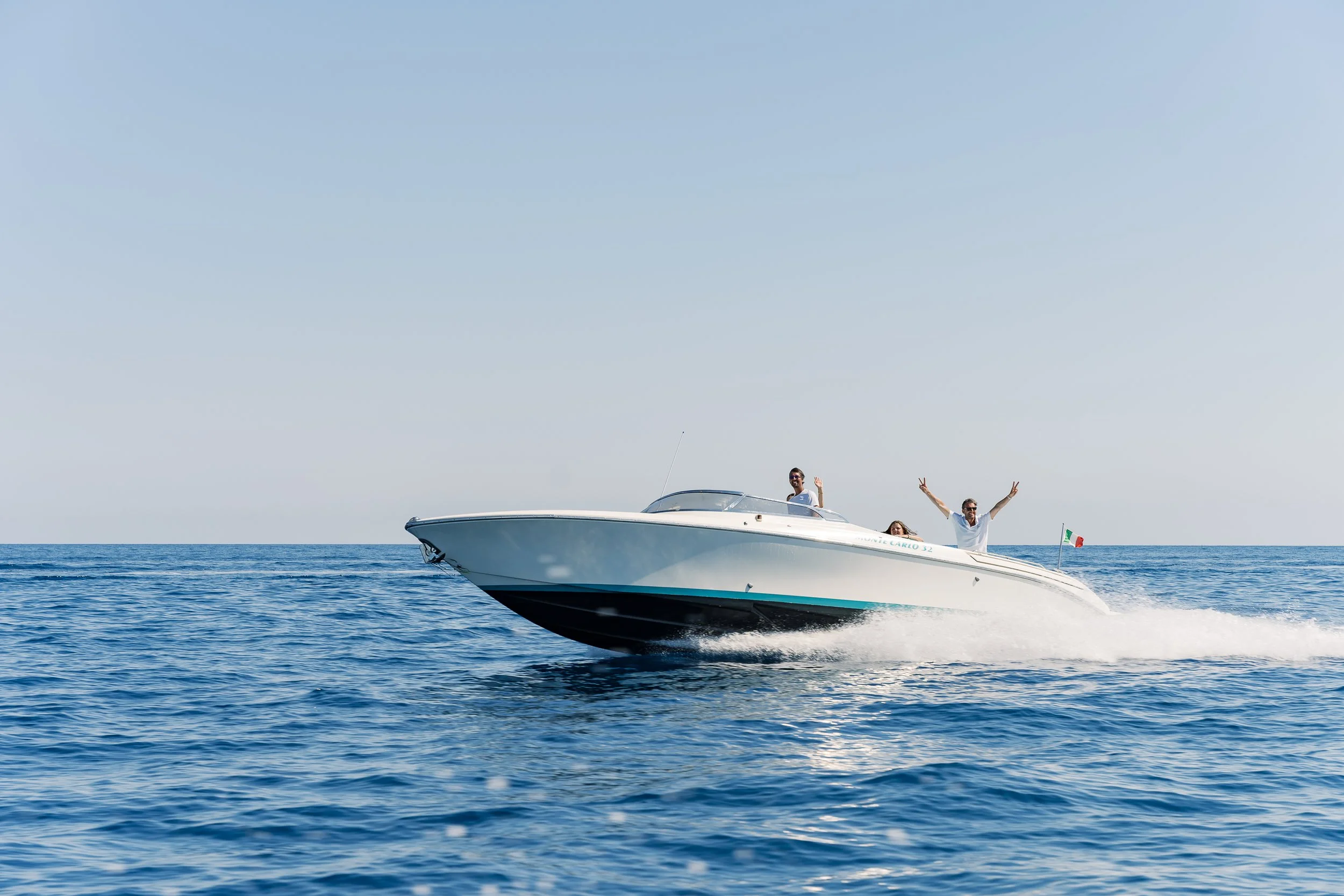 A white speedboat with two people on board, one standing and waving, the other sitting. The boat is moving fast across the water with a small Italian flag at the back. The ocean is calm and the sky is clear.