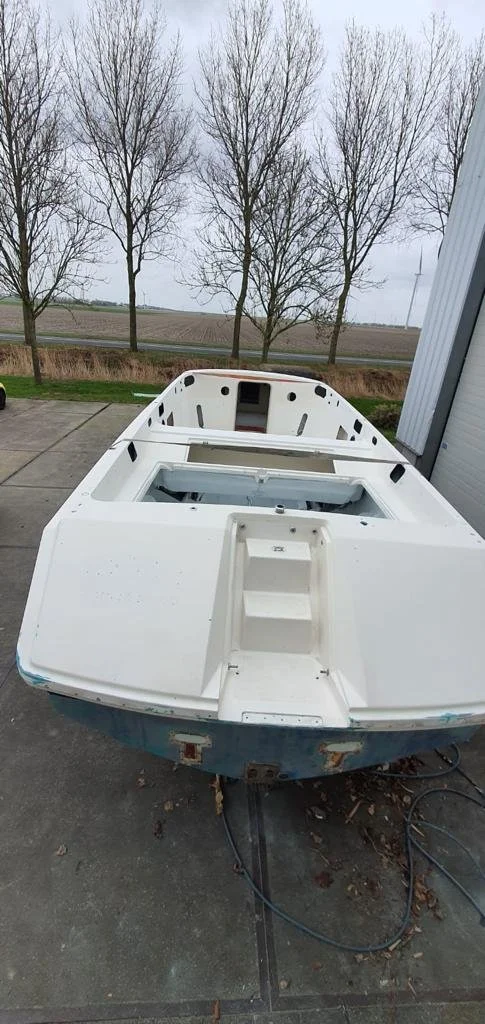 White boat hull on trailer outdoors near a building, with leafless trees and an open field in the background.