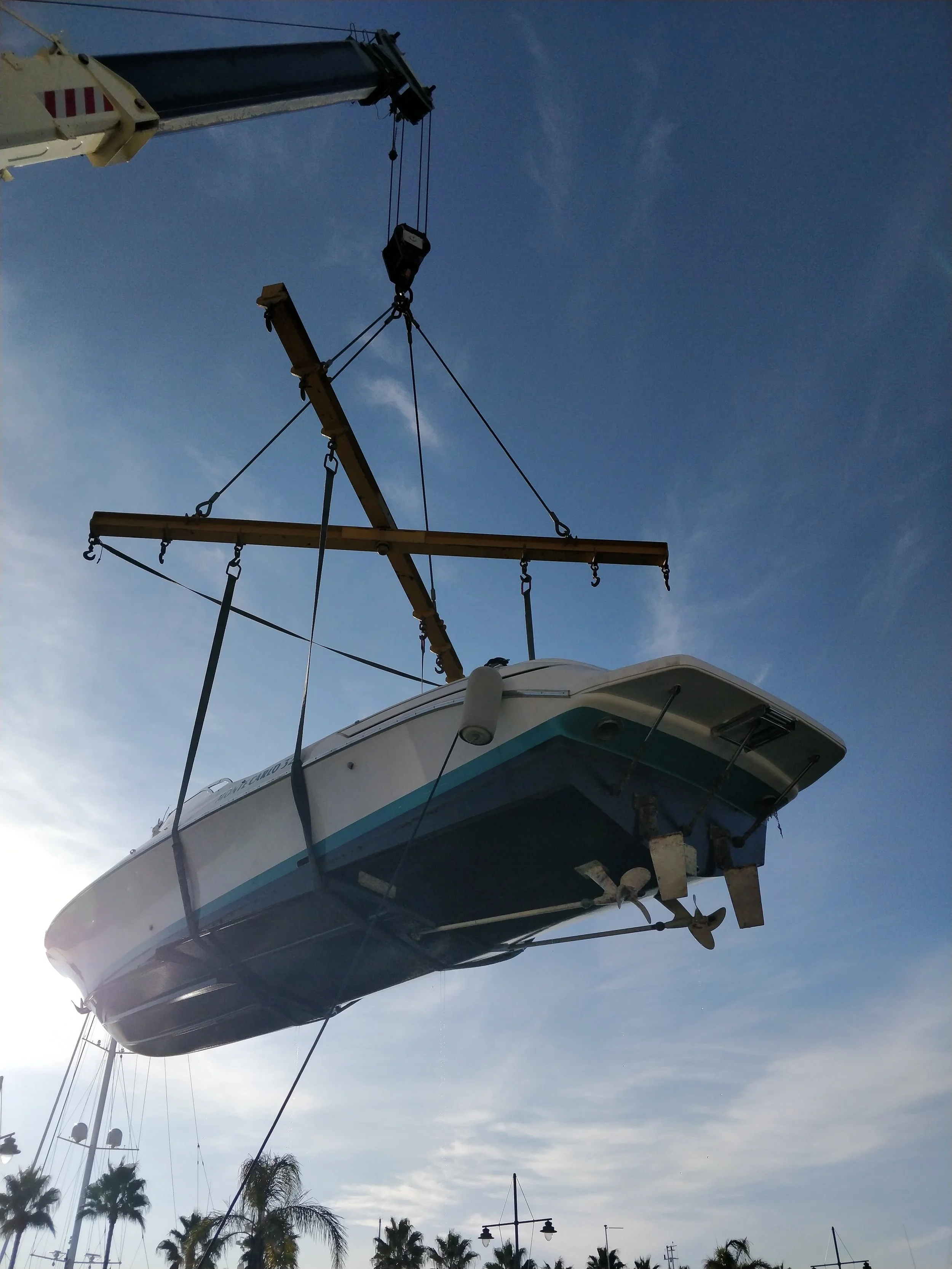 Boat being lifted by a crane against a blue sky with some clouds, with palm trees in the background.