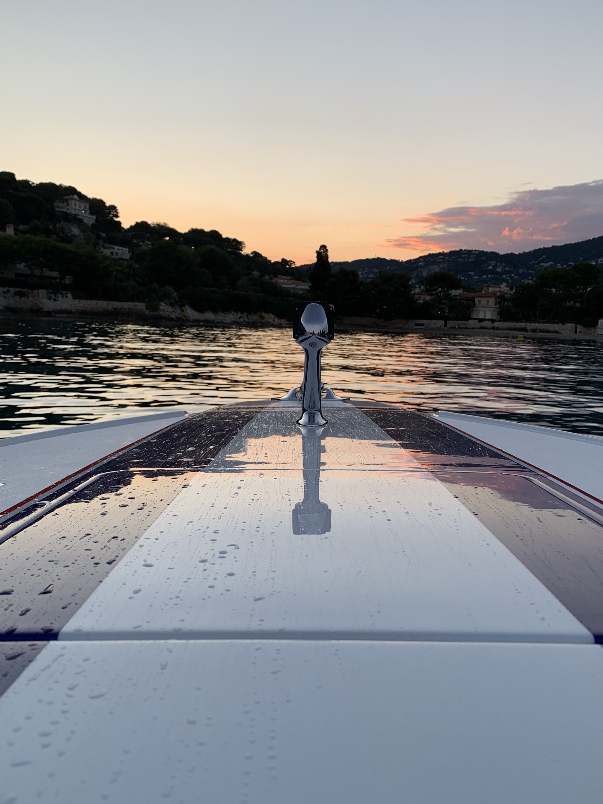 On a boat at sunset, view of water, mountains, and houses in the distance with water droplets on the boat's roof.