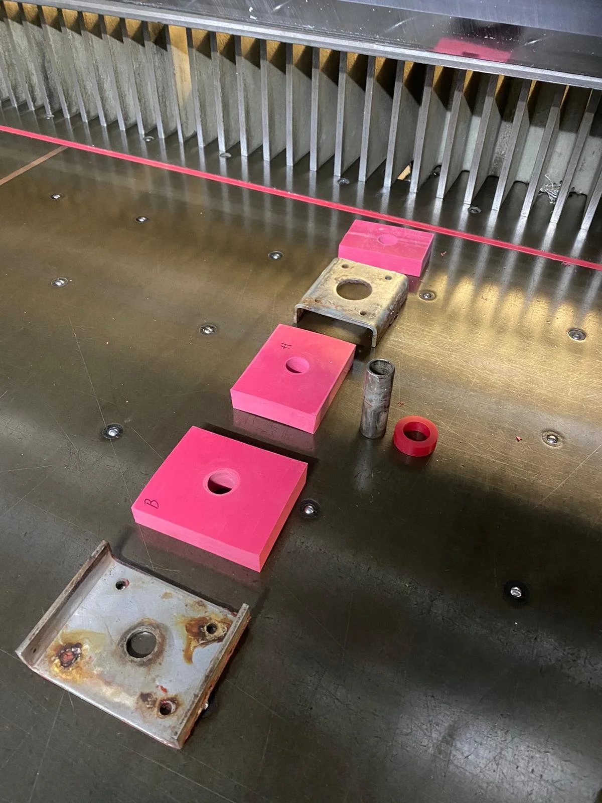 Various metal and plastic components on a metal table, including pink blocks with holes, a rusted metal piece, a metal tube, a small red ring, and a rusted square metal plate with holes. A red line runs across the top of the image.