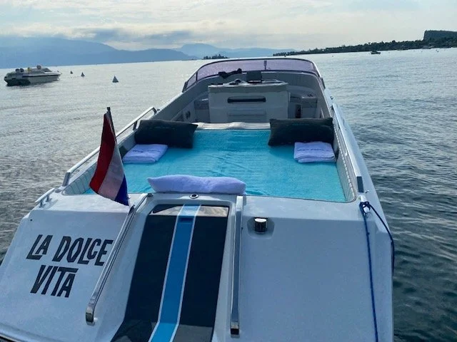 A luxury motorboat on a body of water with a blue cover, towels, and pillows on top, displaying 'La Dolce Vita' on the side, and a flag at the stern, with other boats and distant land in the background.