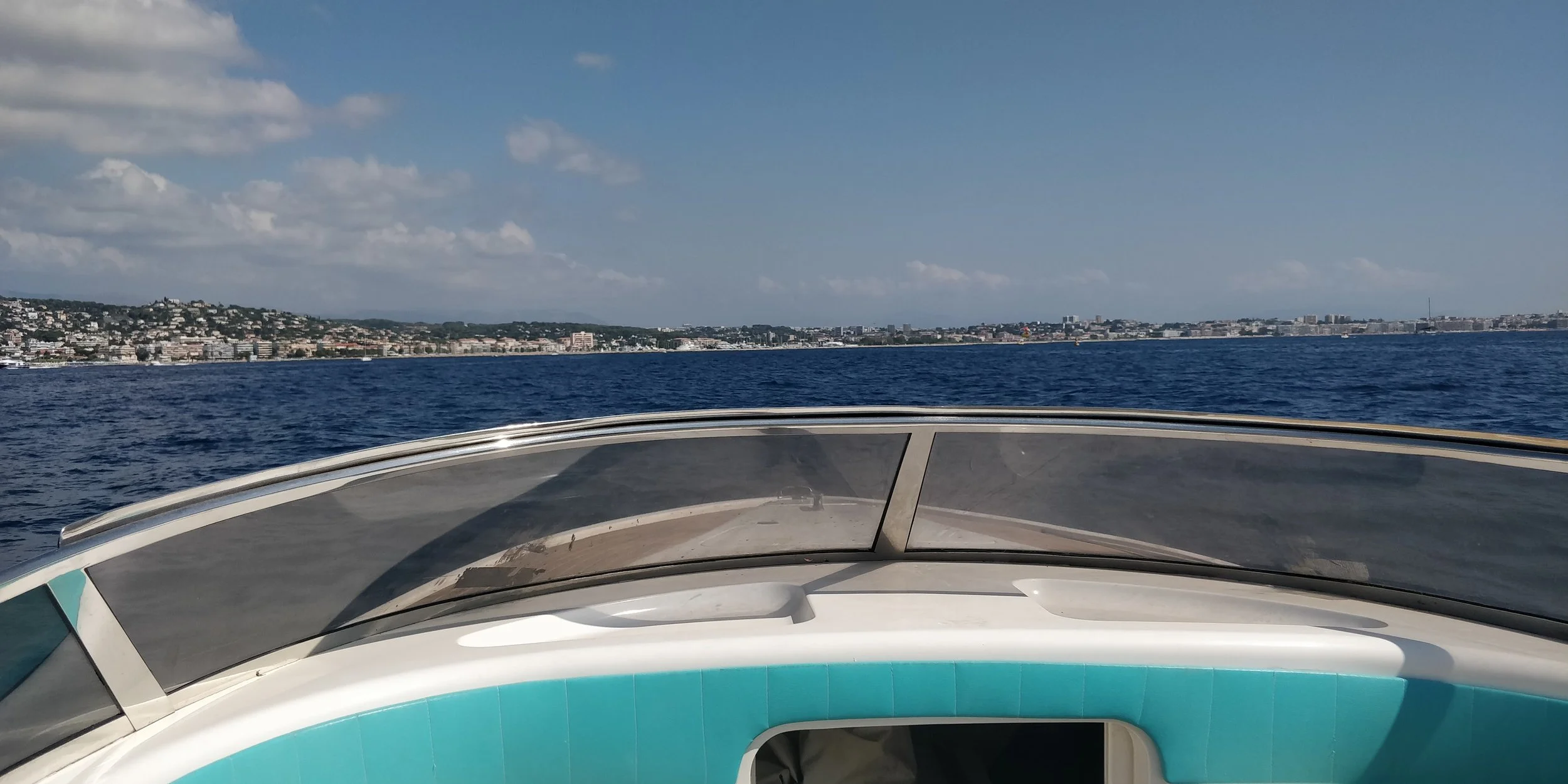 View from a boat showing a cityscape along the coastline, with blue ocean water in the foreground and a partly cloudy sky above.