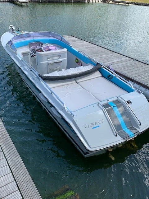 White and blue Speedboat docked at a wooden pier on calm water, with a seating area at the front and a control console at the back.