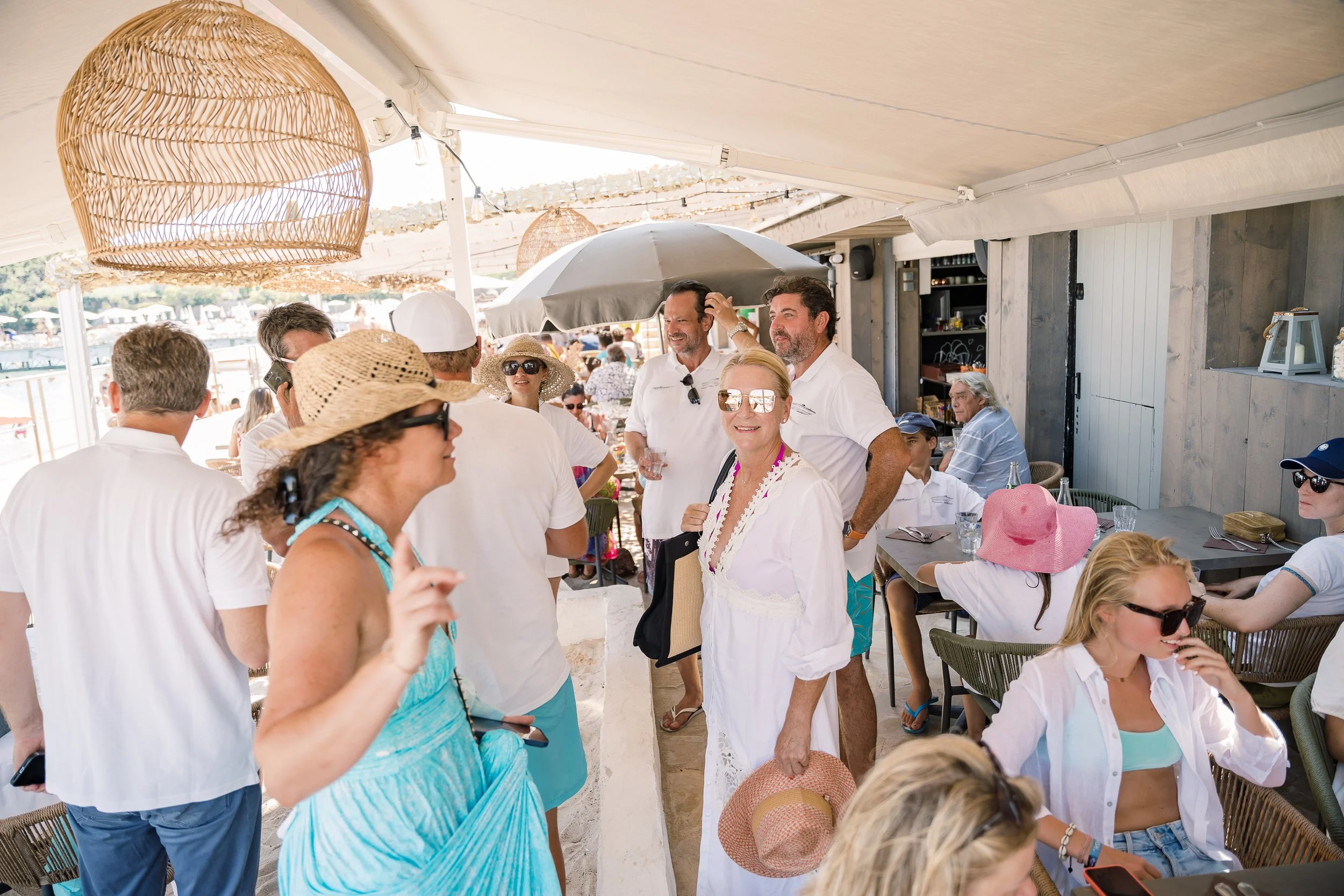 People socializing at an outdoor beachside restaurant under a large tent with hanging wicker light fixtures, some wearing summer hats and sunglasses.