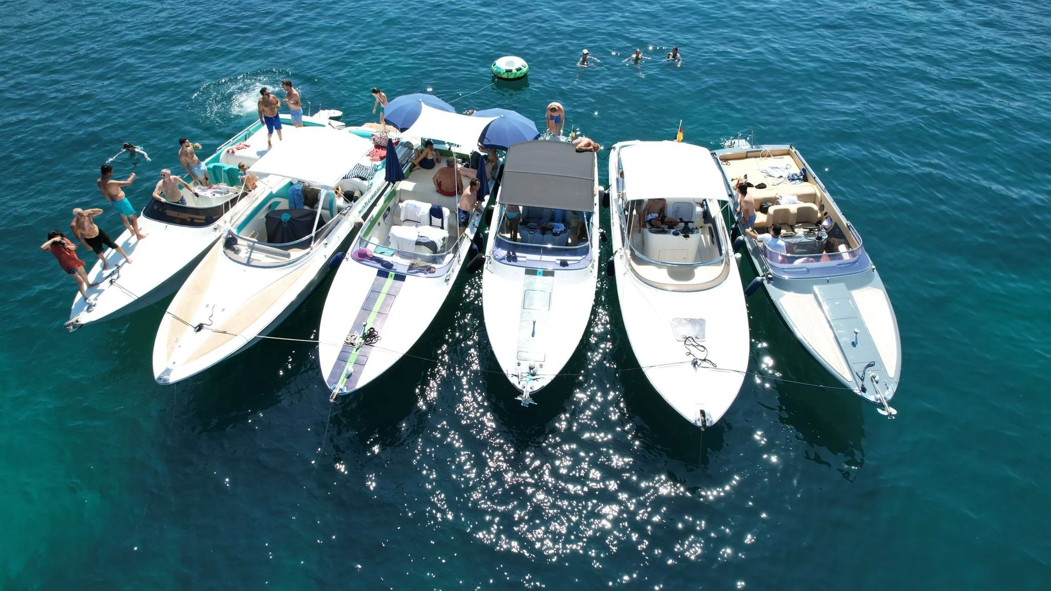 Five boats anchored close together in the water with people swimming and relaxing on them, some under umbrellas, and others on swim floats.