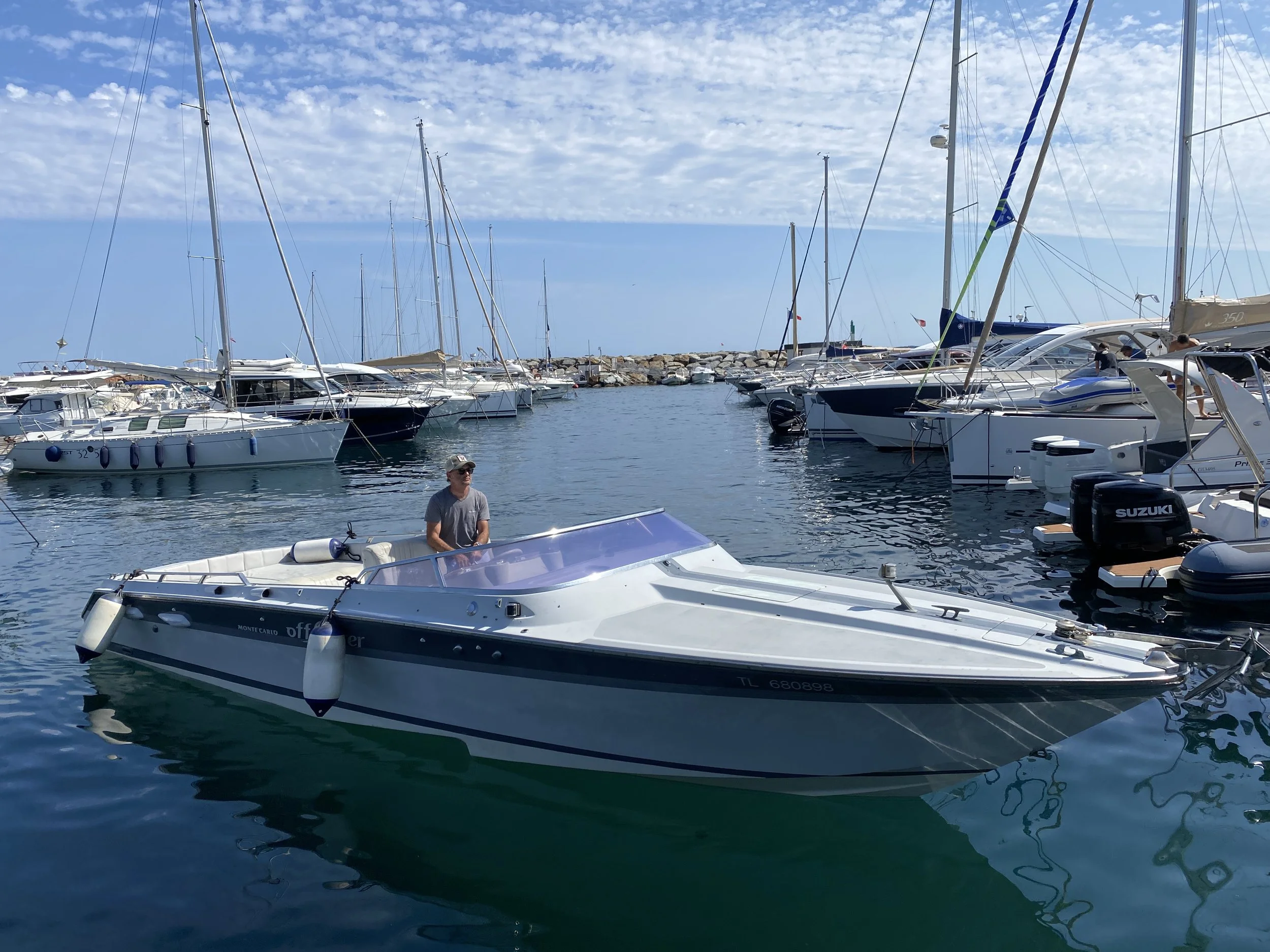 A man standing on a white motorboat in a marina with multiple sailboats and yachts docked nearby on a clear day with scattered clouds.