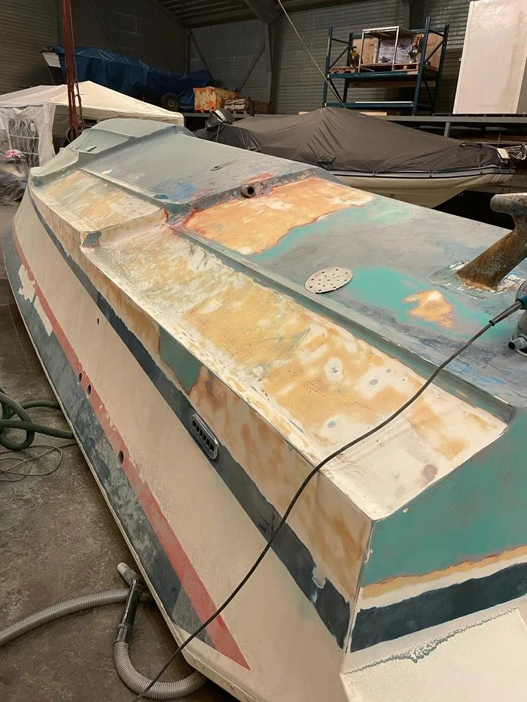 A boat in a workshop undergoing restoration with visible areas of sanding and primer, surrounded by other boats and equipment.