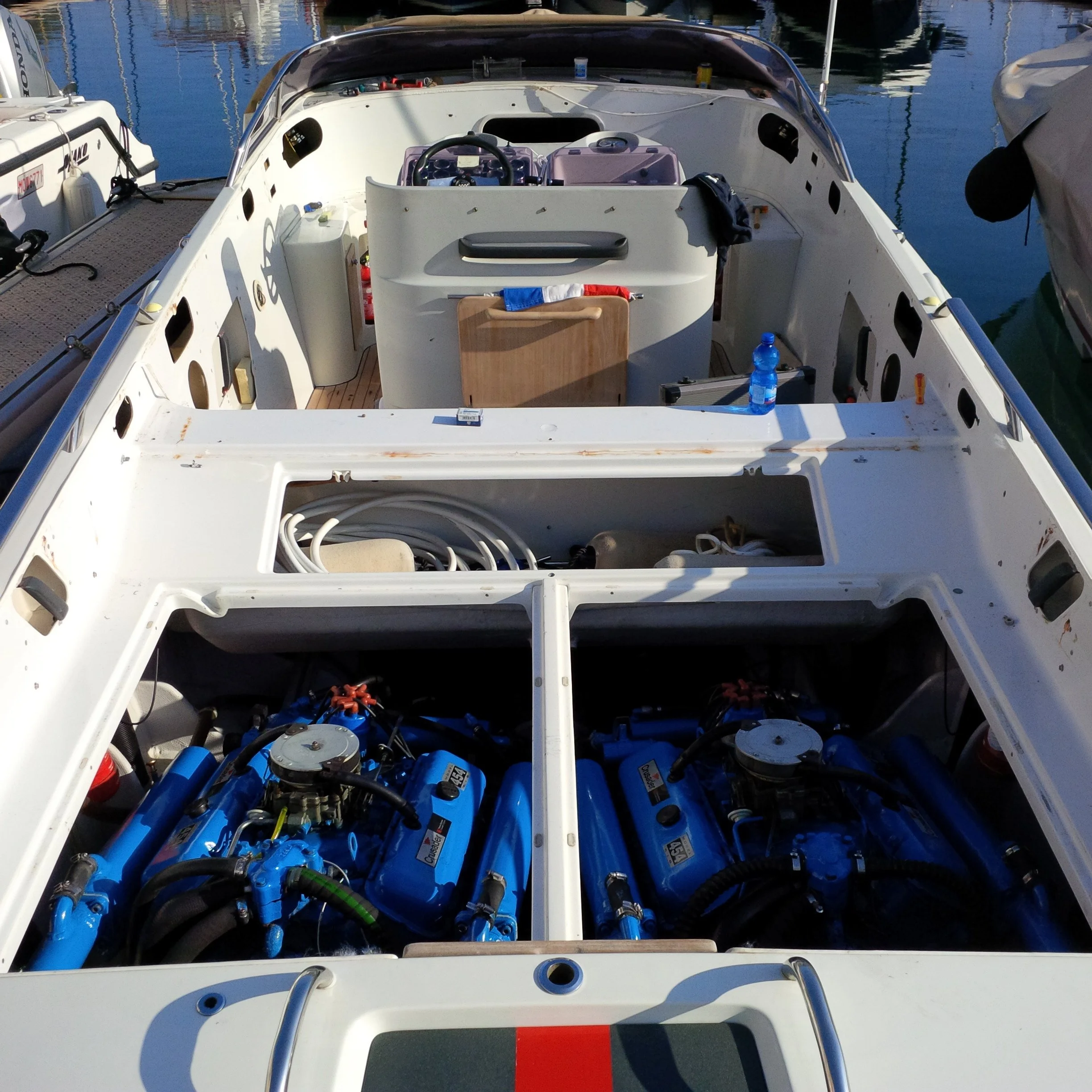 Interior of a white boat with blue engines, storage compartments, and a view of the water and other boats in the background.