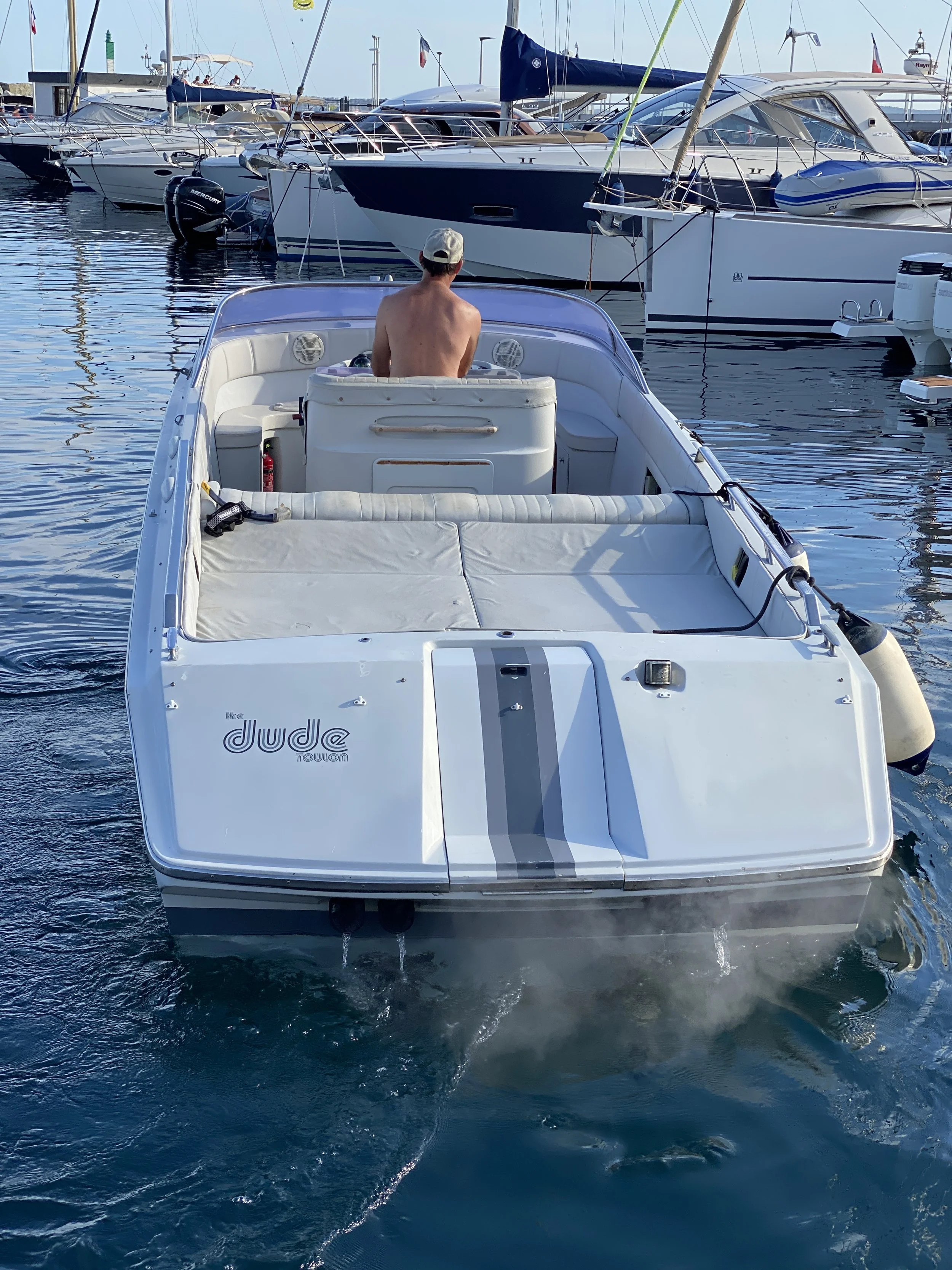 A man sitting shirtless on a white boat named The Jude Toulon, surrounded by several sailboats and motorboats docked at a marina during daytime.