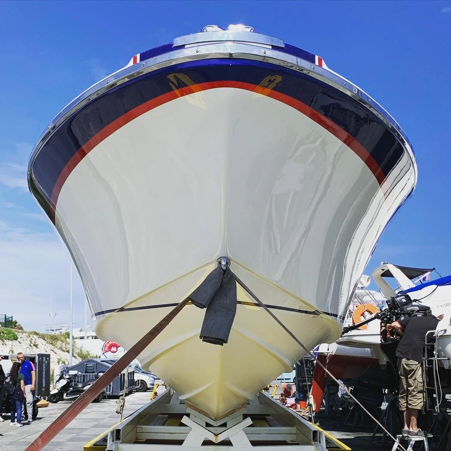 Large white yacht on display at a marina, viewed from beneath, with people and boats in the background.