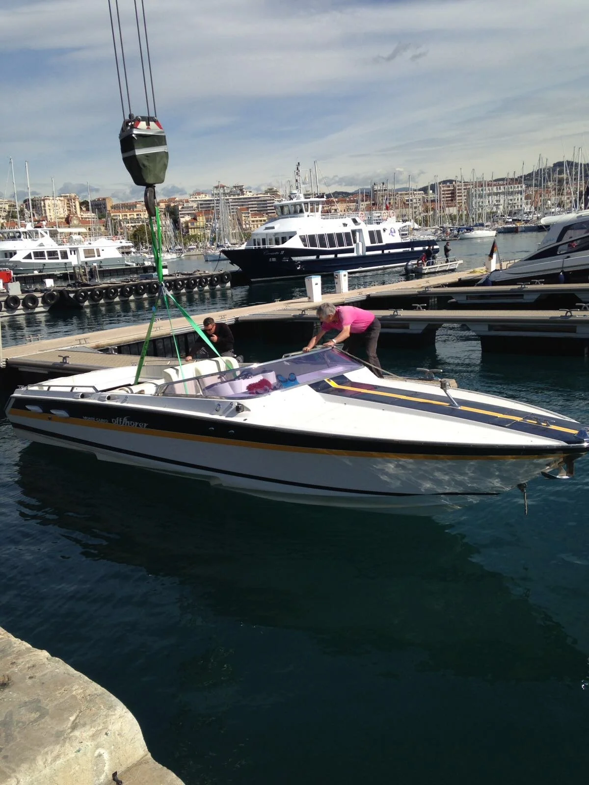 People working on a small white speedboat docked at a marina with various large boats and yachts in the background.