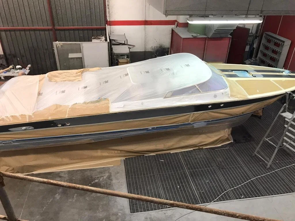 A boat undergoing restoration inside a workshop, covered partially with protective paper and plastic wrap, with tools and equipment around.