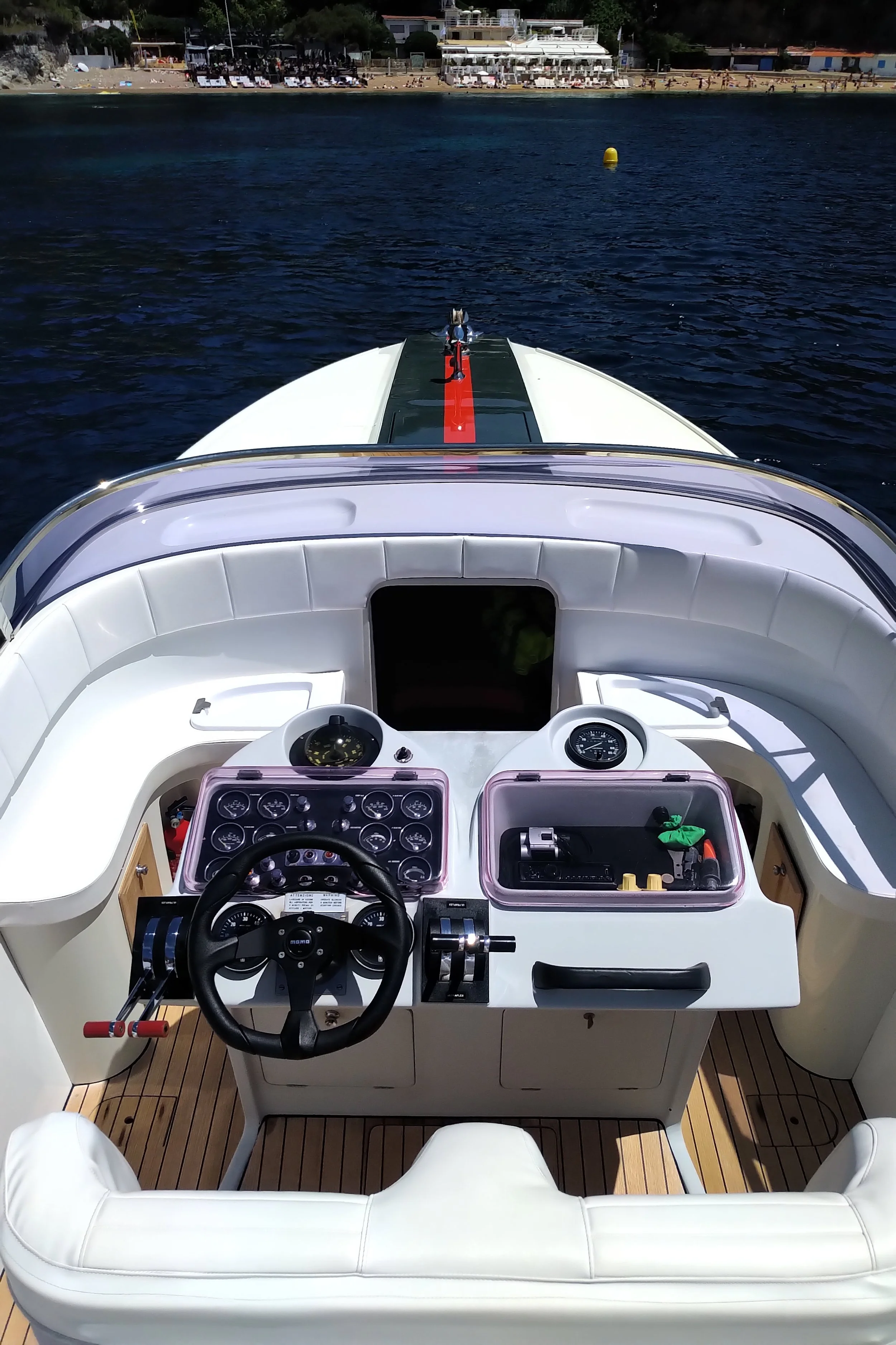 View from the cockpit of a white yacht on the water, showing the control consoles, steering wheel, and seating area, with a shoreline and buildings in the background.