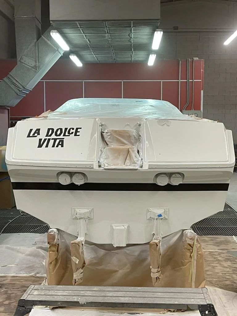 A white racing boat with the words 'LA DOLCE VITA' written on the front, in a workshop or garage, covered in protective tape and paper for painting or finishing.