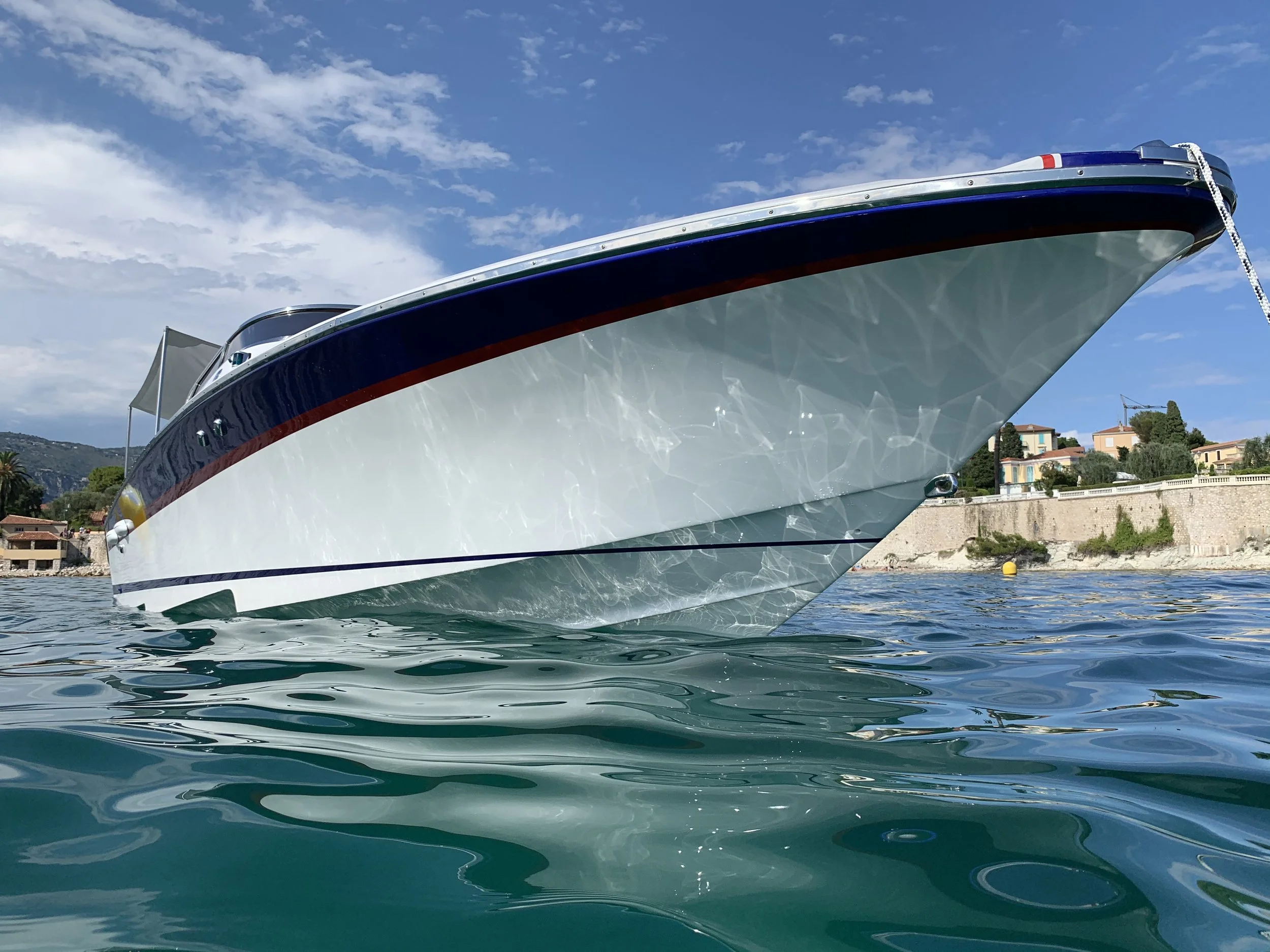 A large white motor yacht floating on calm water near a shoreline with houses, trees, and hills in the background, under a partly cloudy sky.