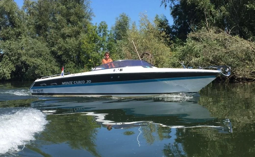 A person is riding on a small white and blue motorboat on a calm river, surrounded by green trees.