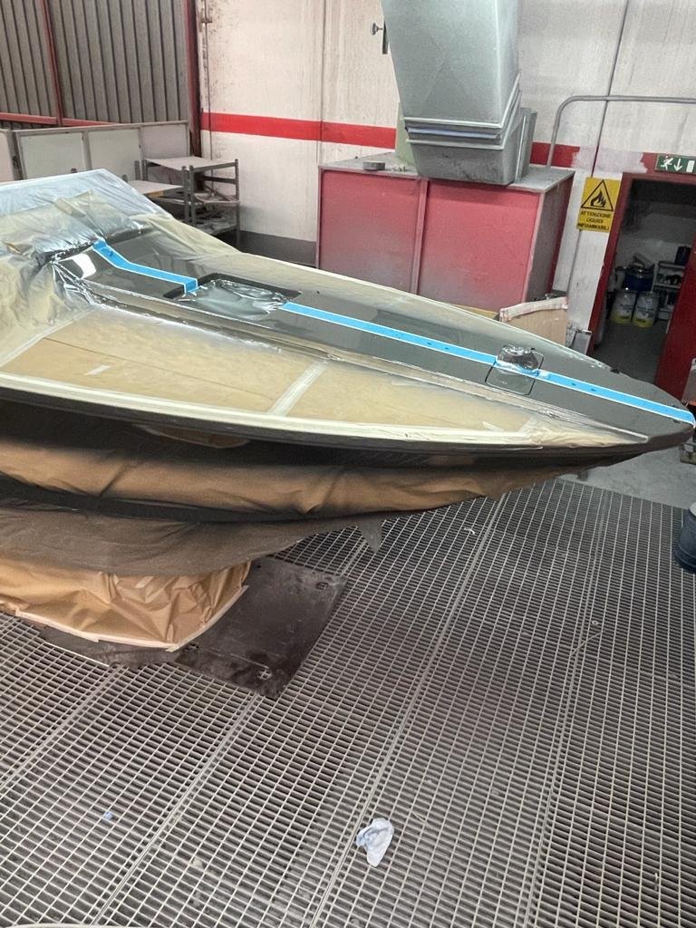The image shows a boat in a workshop or factory setting, with plastic and masking tape applied to parts of the boat, possibly for painting or finishing. There are industrial equipment and shelving in the background.