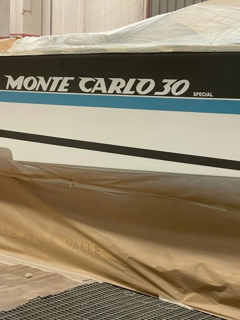 The image shows a boat named 'Monte Carlo 30 Special' in a storage or warehouse area. The boat is partially covered with protective wrapping, and the lower part of the boat is wrapped in brown paper, with part of a metal grate visible at the bottom.