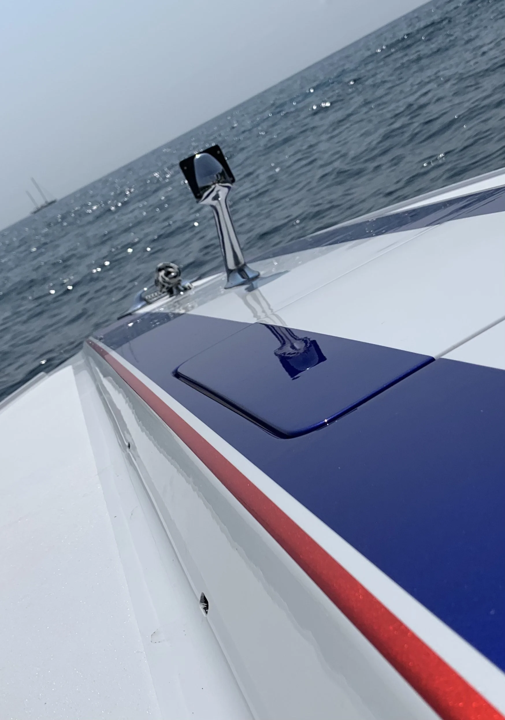 Close-up of a boat's deck with a blue hatch and a mirror mounted on a stand, sailing on the ocean with a distant shoreline.