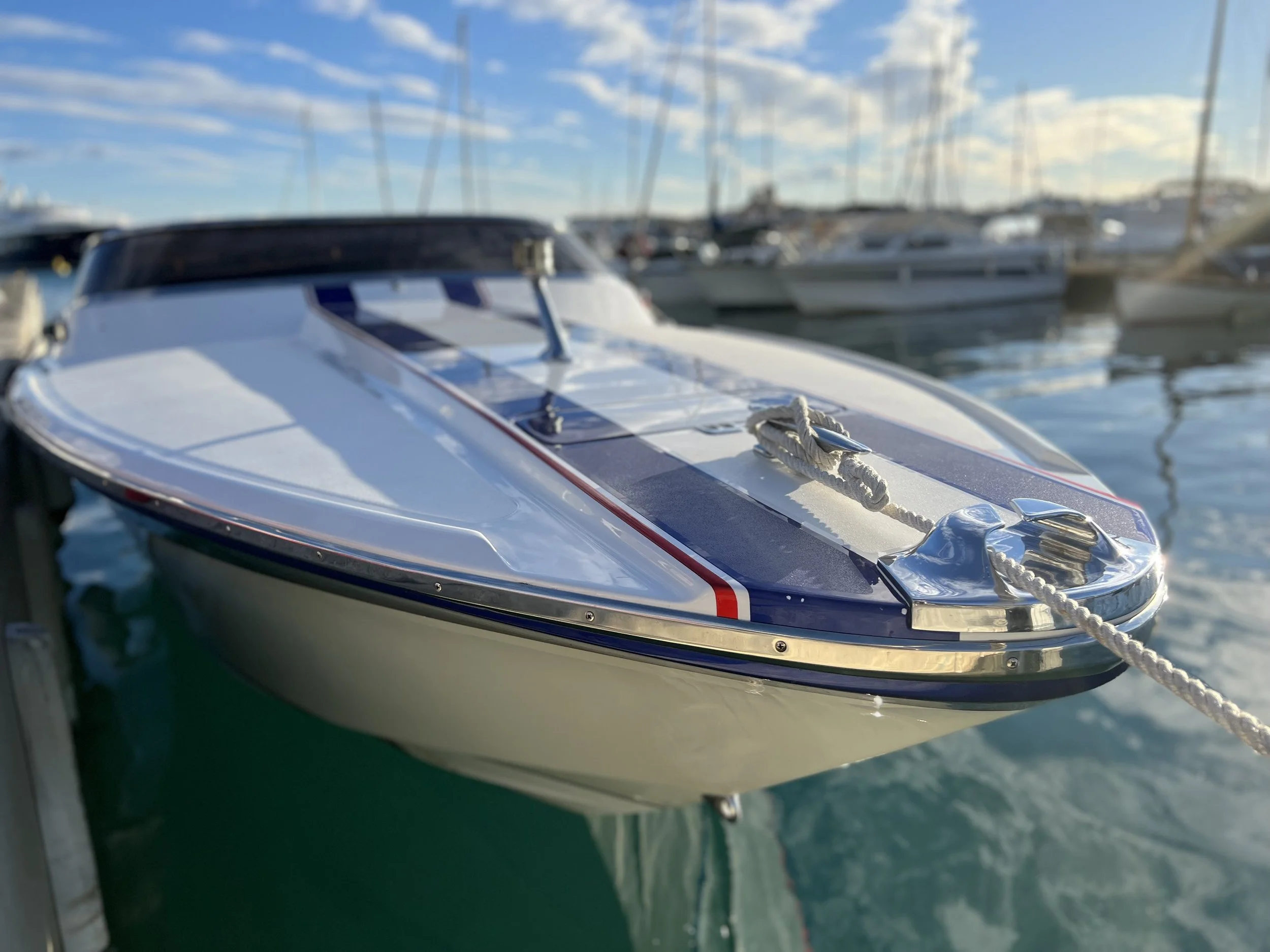 Close-up of a small boat docked at a marina with other boats in the background, under a blue sky with white clouds.