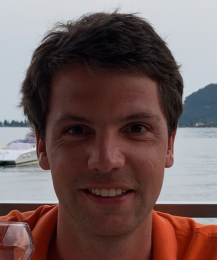 A man smiling, with water and a boat in the background on a cloudy day.