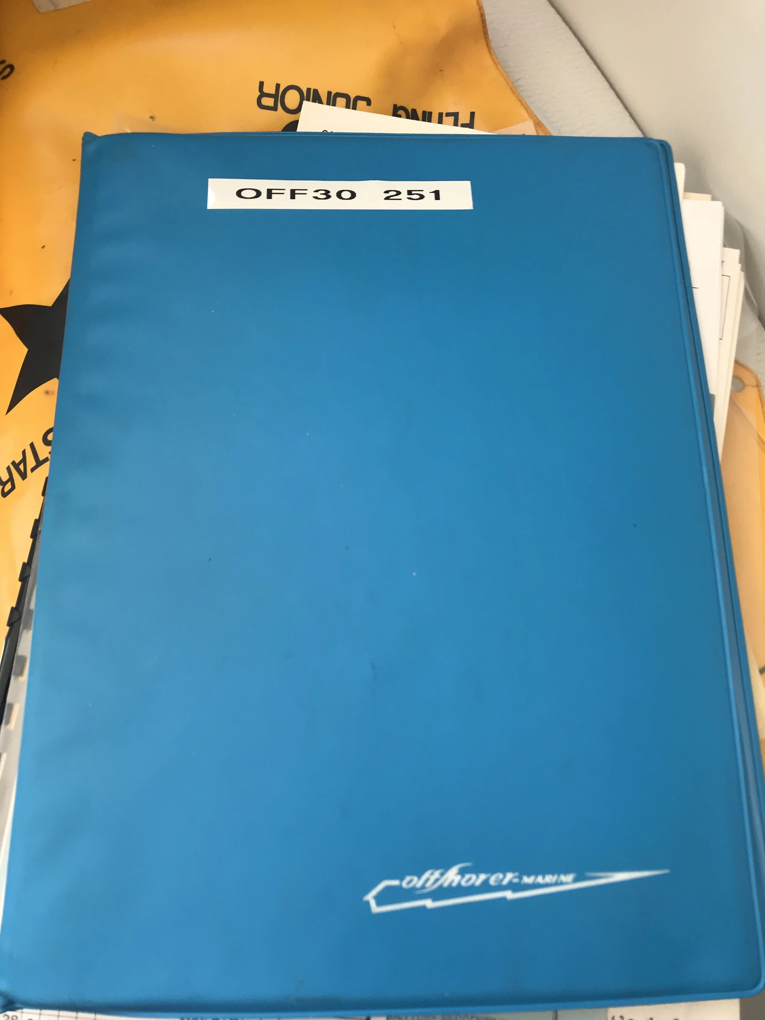 Blue offshores manual or document with the label 'OFF30 251' on the cover, resting on a surface with an orange bag and some papers underneath.